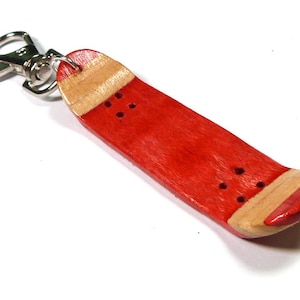 Könnte beinhalten: Ein roter und naturfarbener Skateboard-Schlüsselanhänger mit einem silbernen Metallschlüsselring. Der Schlüsselanhänger ist etwa 10 cm lang.