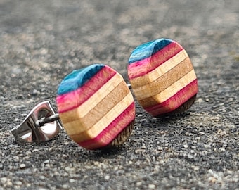 Könnte beinhalten: Ein Paar Holz-Ohrstecker. Jeder Ohrring hat eine abgerundete quadratische Form mit einem gestreiften Design. Die Streifen wechseln sich zwischen natürlichen Holztönen, Rosa und Blau ab. Die Ohrringpfosten sind aus silberfarbenem Metall.