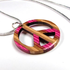 Può includere: Una collana con catena d'argento con un ciondolo a forma di segno della pace in legno e rosa. Il ciondolo è realizzato in legno stratificato con strisce rosa.