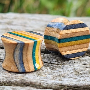 May include: Two wooden ear gauges with a striped pattern in blue, yellow, and green. The gauges are made from recycled skateboard wood.