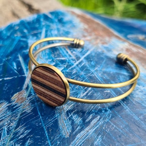 May include: A gold metal bangle bracelet with a round wooden inlay. The inlay has a striped pattern of light and dark wood.
