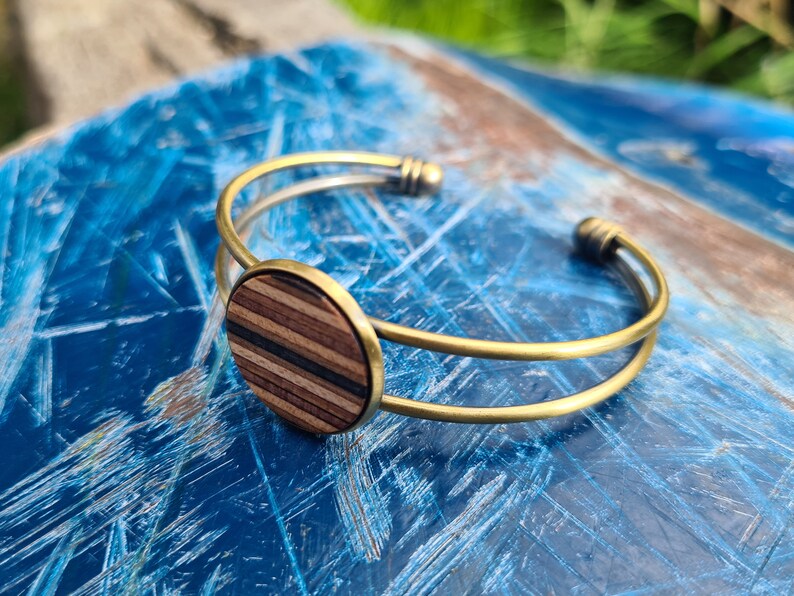 May include: A gold metal bangle bracelet with a round wooden inlay. The inlay has a striped pattern of light and dark wood.
