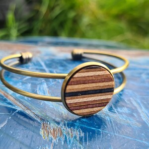 May include: A gold metal bangle bracelet with a round wooden inlay. The inlay has a striped pattern of brown, black, and white wood.