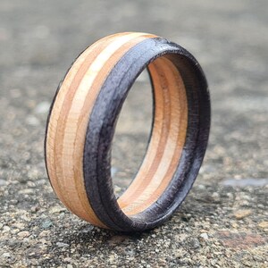 May include: A wooden ring with alternating stripes of light and dark wood, with a black resin inlay. The ring is made of natural materials and has a unique, handcrafted look.