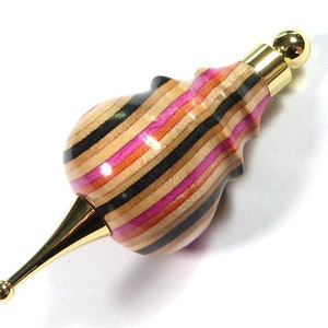 Puede incluir: Un tapón de botella de madera con una parte superior dorada y un patrón a rayas de negro, marrón, rosa y naranja.