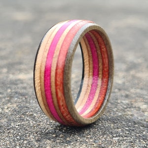 May include: A wooden ring with pink, red, and brown stripes. The ring is made from recycled skateboard wood.