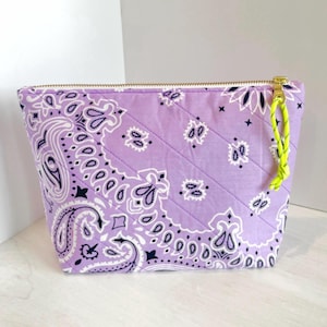 May include: A quilted lavender cosmetic bag with a gold zipper and a neon yellow pull. The bag features a paisley pattern in white and black. The bag is rectangular with a flat bottom.