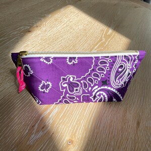 May include: A quilted purple pouch with a white paisley pattern. The pouch has a gold zipper and a pink pull tab. The bag is rectangular with a trapezoid shape and is sitting on a wooden surface.