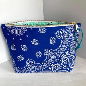 May include: A blue bandana-print zippered pouch with a gold zipper and a teal pull. The pouch features a white paisley design on a blue background. The interior lining is a light green color.