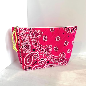 May include: A hot pink quilted cosmetic bag with a paisley bandana print in white and black. The bag has a gold zipper and a yellow pull cord. The bag is sitting on a white surface.
