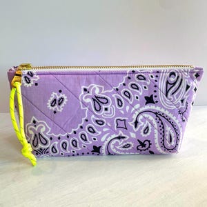 May include: A lavender quilted cosmetic bag with a gold zipper and a neon yellow pull. The bag features a paisley pattern in white and black. The bag is rectangular with a trapezoid shape.