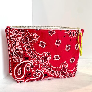May include: A red bandana-print zippered pouch with a gold-colored zipper and a yellow pull cord. The pouch features a quilted pattern and a classic paisley design in white and black.