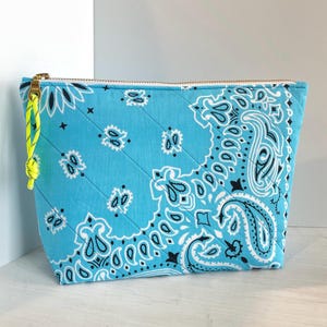 May include: A quilted, rectangular cosmetic bag with a light blue bandana print. The bag features a gold zipper and a neon yellow pull. The bandana print includes white and black paisley designs. The bag is a stylish accessory for storing makeup or toiletries.