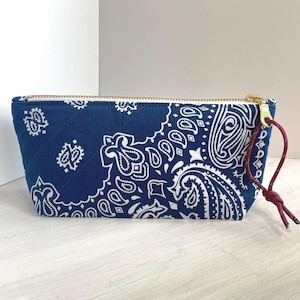 May include: A navy blue quilted cosmetic bag with a white paisley pattern. The bag has a gold zipper and a red cord loop. The bag is rectangular and sits on a light-colored surface.