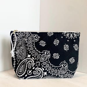 May include: Black and white paisley print cosmetic bag with a gold zipper and white cord pull. The quilted bag features a classic bandana design, offering a stylish storage solution for makeup and toiletries.