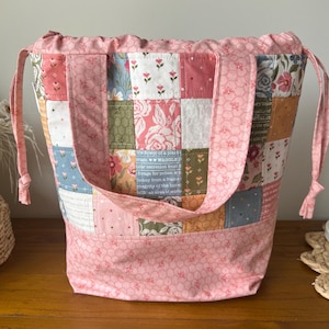 May include: A patchwork tote bag with a pink floral print base and handles. The body of the bag is made of various fabric squares in pastel colours and floral patterns. The bag has a drawstring closure and is on a wooden surface.
