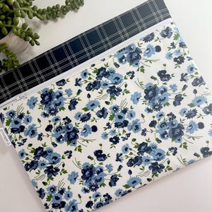 May include: A zippered pouch with a floral pattern and a plaid pattern. The floral pattern features blue flowers on a white background. The plaid pattern is black and navy blue. The pouch has a white zipper and a small tag that says "RED RASPBERRY CO."