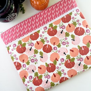 May include: A rectangular zippered pouch featuring a white background with a pattern of pumpkins in shades of red, orange, and pink, interspersed with purple flowers and green leaves. The top border is pink with the word "boo" repeated. The zipper is white.