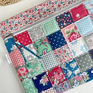 May include: A rectangular fabric pouch featuring a patchwork design of various floral, cat, and geometric patterns in shades of pink, blue, green, and grey. The top section has a floral border and a silver zip with a blue fabric pull tab.