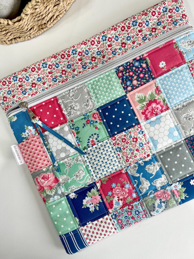 May include: A rectangular fabric pouch featuring a patchwork design of various floral, cat, and geometric patterns in shades of pink, blue, green, and grey. The top section has a floral border and a silver zip with a blue fabric pull tab.