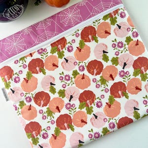 May include: A zippered pouch with a white background, featuring a pattern of pumpkins in shades of pink, peach, and red, interspersed with small purple flowers and green leaves. The top border is a solid pink with a white spiderweb design.