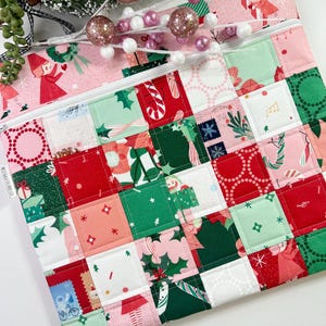 May include: A quilted Christmas-themed pouch with a white zipper. The pouch features a patchwork design with red, green, and pink squares, each showcasing festive patterns like candy canes, elves, and snowflakes. The top section has a pink background with elf illustrations.