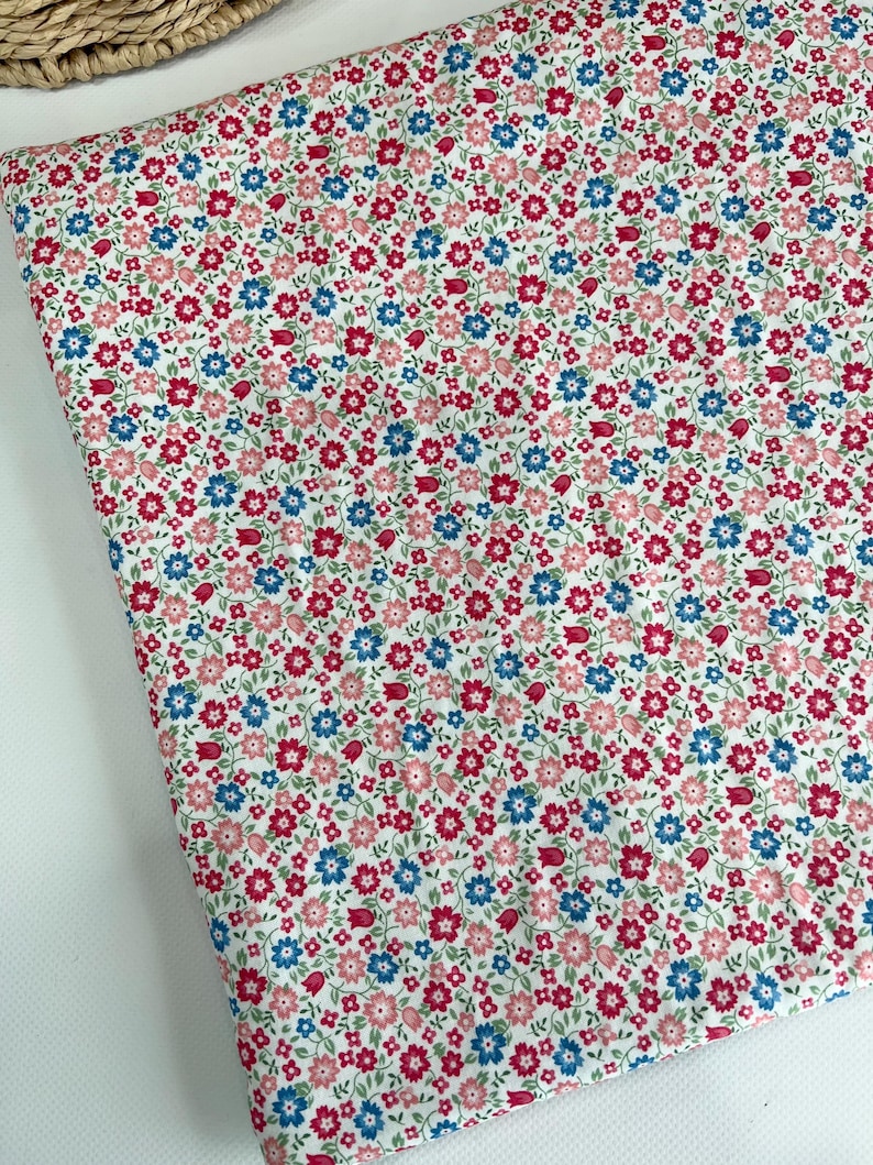May include: A folded piece of fabric featuring a dense, repeating pattern of small flowers in shades of pink, red, and blue on a white background. The floral design includes tiny blossoms and buds with green stems scattered across the surface.