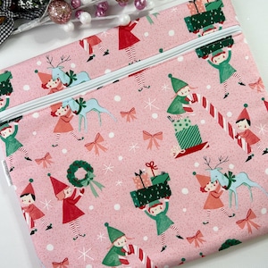May include: Pink Christmas-themed zippered pouch featuring a repeating pattern of elves, reindeer, gifts, and snowflakes. The elves are dressed in green and red, carrying presents and candy canes. The pouch has a white zipper.