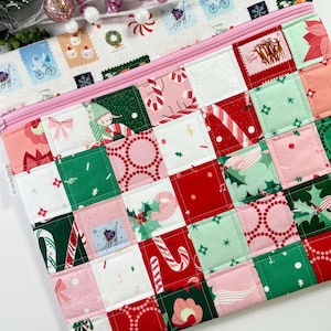 May include: A quilted Christmas-themed zippered pouch with a patchwork design. The pouch features squares of fabric with holiday patterns like candy canes, stamps, and elves. The zipper is pink, and the top section has a white background with stamps.