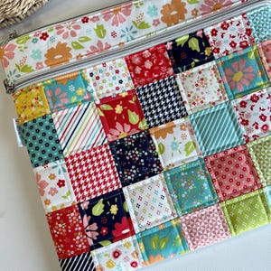 May include: A quilted patchwork zippered pouch featuring a variety of colorful fabric squares. The squares include floral, striped, and patterned designs in shades of red, blue, yellow, and green. The top border and zipper area have a floral pattern on a white background.