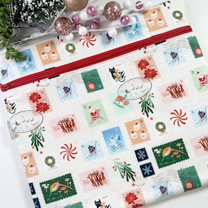 May include: White zippered pouch with a festive stamp design featuring cats, reindeer, and Santa Claus. The stamps are in various colors, with a red zipper. Decorative holiday accents are in the background.