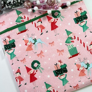 May include: Pink Christmas-themed fabric with a repeating pattern of elves, reindeer, gifts, and bows. The elves are dressed in red and green outfits. A green zipper runs across the top of the fabric.
