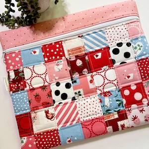 May include: A quilted zipper pouch with a patchwork design. The pouch features squares of red, pink, white, and blue fabric with various patterns, including polka dots, stripes, and hearts. A white zipper and pink polka dot fabric trim the top.