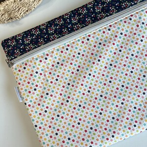 May include: A fabric zipper pouch with a white background covered in small, colorful flowers. The top border is navy blue with a floral pattern. A silver zipper runs across the top. The pouch is likely used for storage or organization.