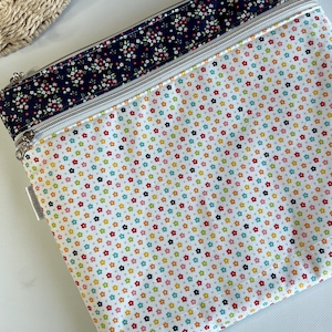 May include: A white zippered pouch with a colorful floral pattern. The pouch features a navy blue floral border and a silver zipper. The main body of the pouch is covered in small, multi-colored flowers.