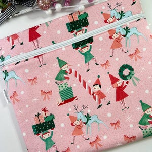 May include: A pink zippered pouch with a festive Christmas pattern. The design features elves, reindeer, gifts, and bows. The pouch has a white zipper and is decorated with white snowflakes.