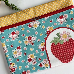 May include: A zipped pouch featuring a teal fabric with a red, yellow, and white floral pattern. The top border is yellow with a white grid design. A white circular patch on the right side displays a red plaid strawberry with a flower.