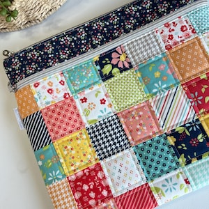 May include: A quilted patchwork zipper pouch with a variety of floral, striped, and patterned fabric squares in shades of red, orange, yellow, blue, and green. The top border is navy blue with a small floral print. A silver zipper runs along the top.