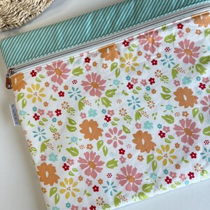 May include: A floral print zippered pouch with a white background and colorful flowers in shades of pink, orange, yellow, and blue. The top edge features a mint green and white striped fabric. A small tag reads "Wild Raspberry Co."