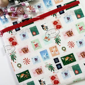 May include: A white fabric pouch with a red zipper, covered in colorful postage stamp designs. The stamps feature holiday-themed illustrations, including cats, Santa, and reindeer. Green sprigs and red accents complete the festive look.