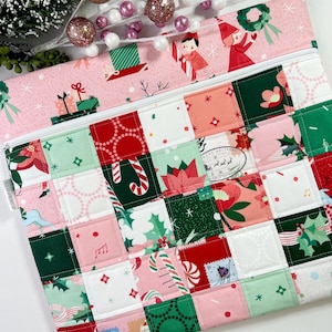 May include: A quilted Christmas-themed zippered pouch with a patchwork design. The pouch features a pink top with illustrations of elves and gifts, and a patchwork of red, green, and pink squares with holiday patterns like candy canes and holly. A white zipper runs across the top.