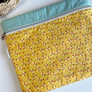 May include: A yellow floral print zippered pouch with a teal and white striped top. The pouch features a silver zipper and a small white tag. The floral pattern includes red, white, and yellow flowers.