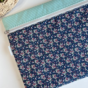 May include: A navy blue zippered pouch with a floral pattern of small pink, white, and yellow flowers. The top section is a light green with white stripes. The pouch has a silver zipper and a small white tag.