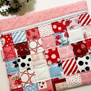 May include: A quilted zippered pouch with a patchwork design. The patchwork features squares with strawberries, polka dots, stripes, and hearts in red, pink, and white. The top and zipper are pink with a white zipper.