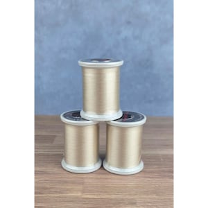 May include: Three spools of metallic gold embroidery thread stacked in a pyramid formation. Each spool features a white plastic core with a black label on the top edge, containing white text that reads Superior Threads and So Fine.