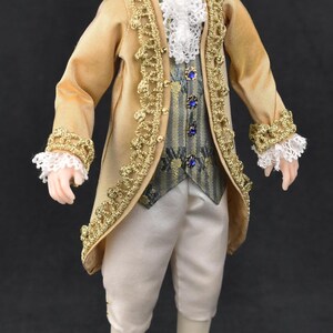 May include: A doll with white hair wearing a gold coat with gold trim, a white lace cravat, a grey patterned vest with blue buttons, and white breeches. Brown shoes with gold buckles are worn. Text reads: Example of Dressed Doll Using Pattern-Doll Is Not Included.