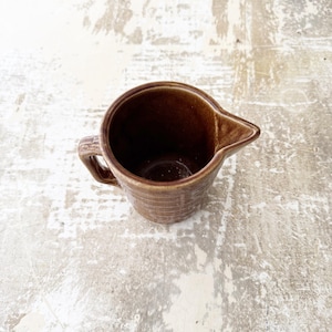 以下が含まれることがあります： 取っ手と注ぎ口が付いた茶色のセラミック製のクリーマー。クリーマーは表面がテクスチャ加工されており、空です。
