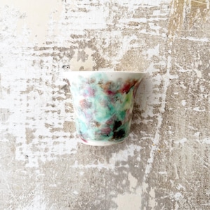 May include: A small, white ceramic cup with a unique, abstract design featuring shades of green, pink, and brown. The cup is ideal for serving coffee, tea, or other beverages.