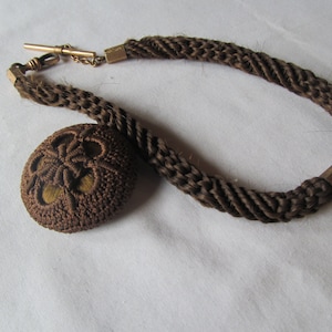 May include: A brown braided hair chain with a gold clasp and a brown hair fob. The fob is a round, woven design with a hole in the center.
