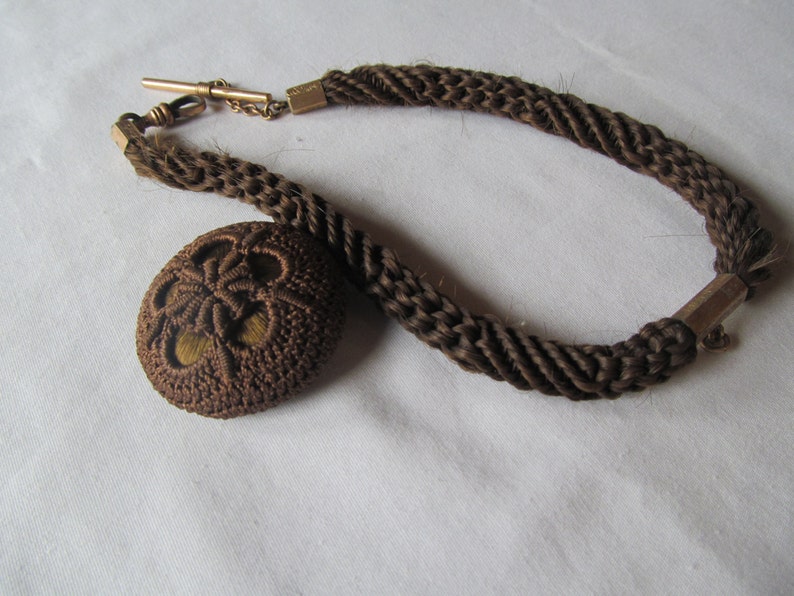 May include: A brown braided hair chain with a gold clasp and a brown hair fob. The fob is a round, woven design with a hole in the center.